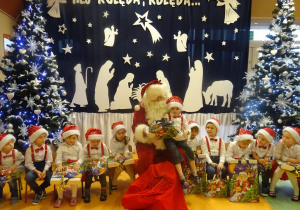 Chłopiec siedzi na kolanach Mikołaja, ogląda prezent. Wokół siedzi grupa dzieci.